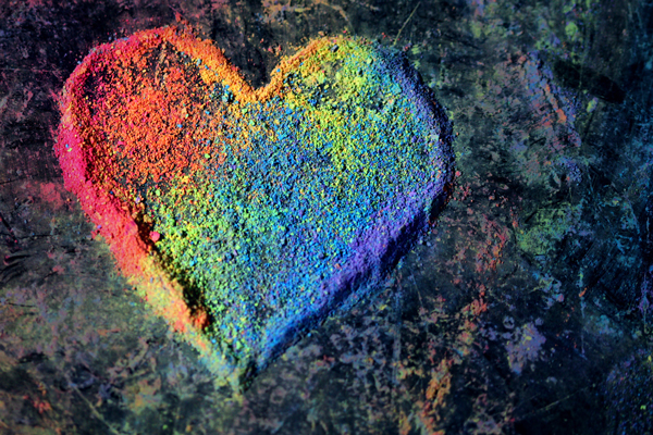 A photo of a heart shape composed of rainbow-colored chalk dust on a dark, grey background.