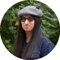 Sharlotte, a person with long dark hair and dark glasses is wearing a gray hat and a jean jacket and is looking at the camera