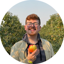 Eóin smiles at the camera while holding an apple below their face